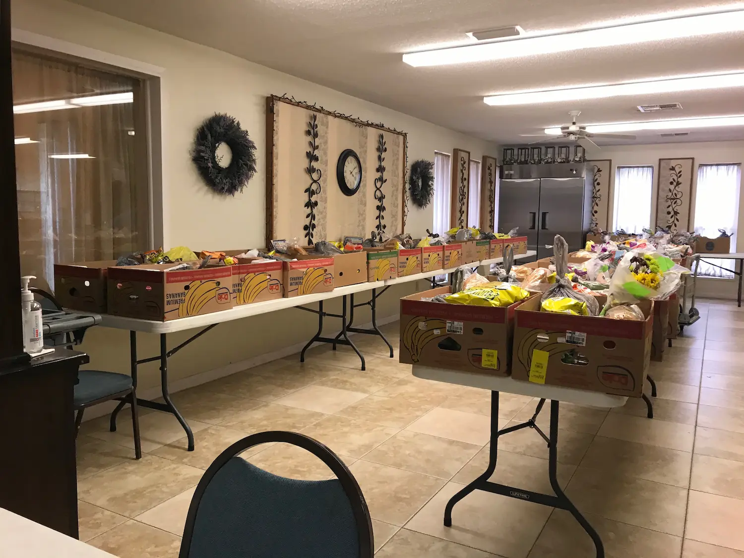 A room with long tables lined with boxes filled with assorted groceries like bread and snacks, aiming to bridge the gap in local hunger. Chairs surround the tables, and wreaths adorn the walls. A clock is visible, and large windows allow natural light to flood in.