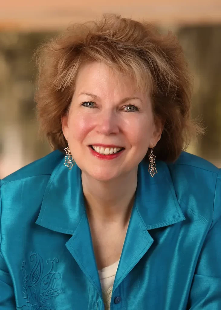 A smiling woman with short, light brown hair wears a teal blouse with embroidered details and dangling earrings, embodying strength and recovery, photographed against a softly blurred background.