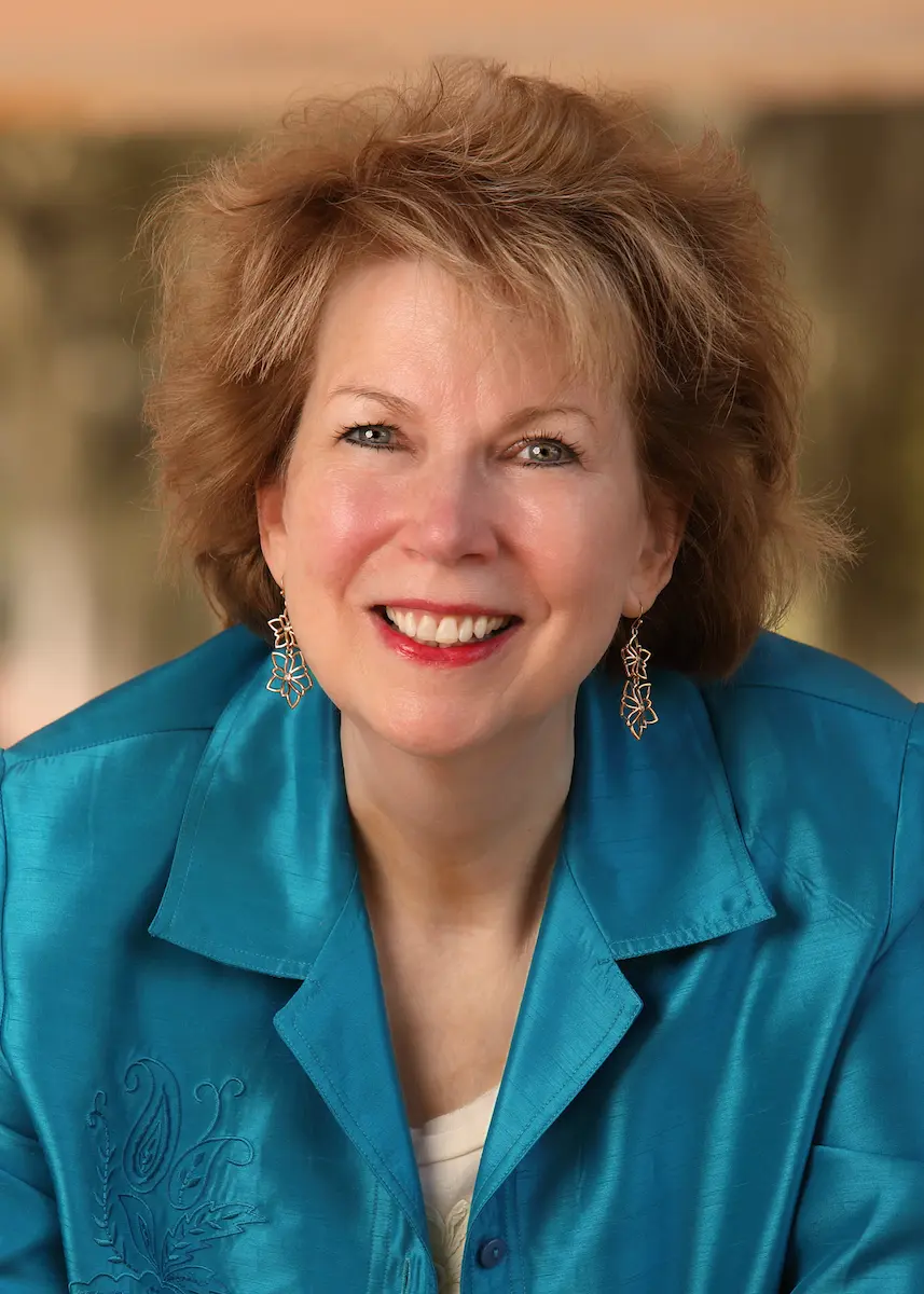 A smiling woman with short, light brown hair wears a teal blouse with embroidered details and dangling earrings, embodying strength and recovery, photographed against a softly blurred background.