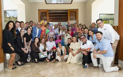 A large group of people, some in colorful costumes and others in business or casual attire, smile at the camera on a marble floor in a well-lit room at Fairmont Mayakoba, supporting Make-A-Wish Mexico.