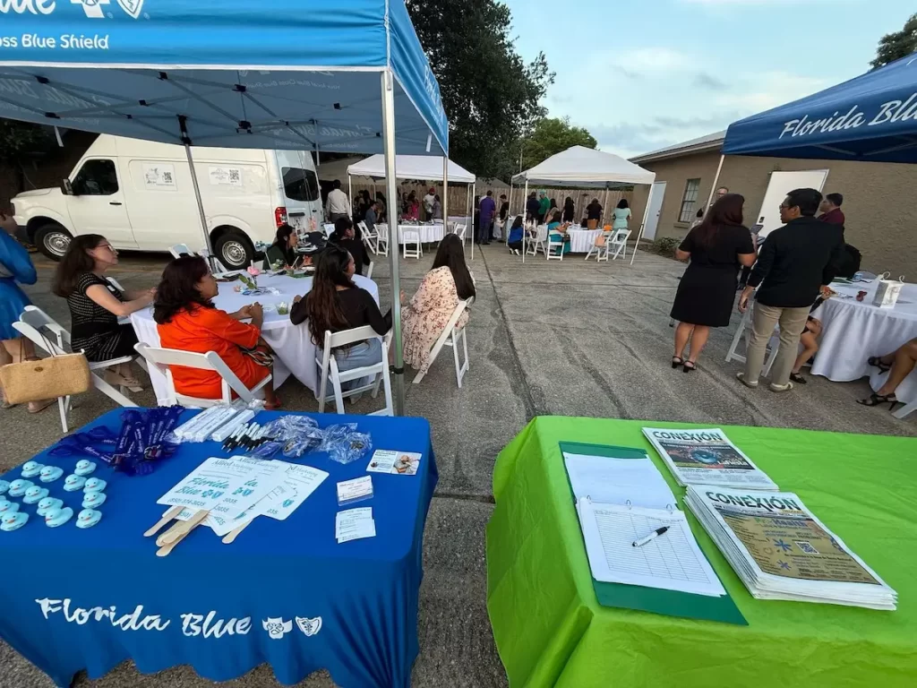 Several people are seated and standing under white tents at an outdoor event. In the foreground, tables with brochures, pens, and promotional items are covered with blue and green cloths labeled “Florida Blue.”.