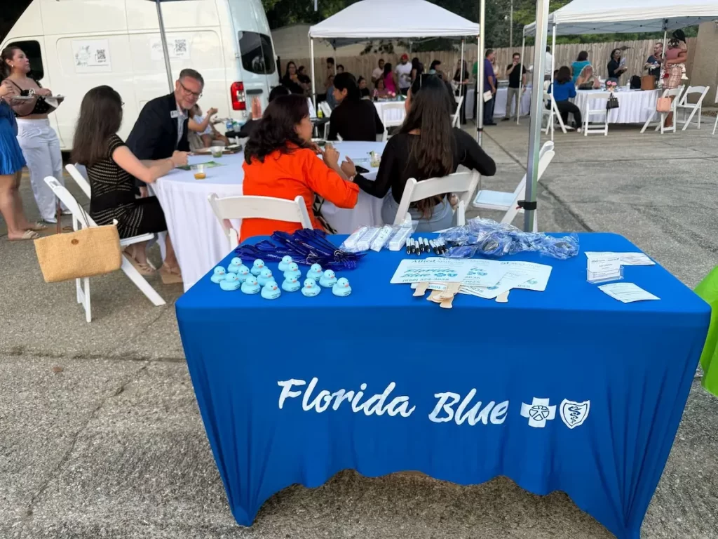 A blue tablecloth with "Florida Blue" logos covers a table displaying rubber ducks, pens, and brochures at an outdoor event. People sit and talk at tables under canopies in the background.