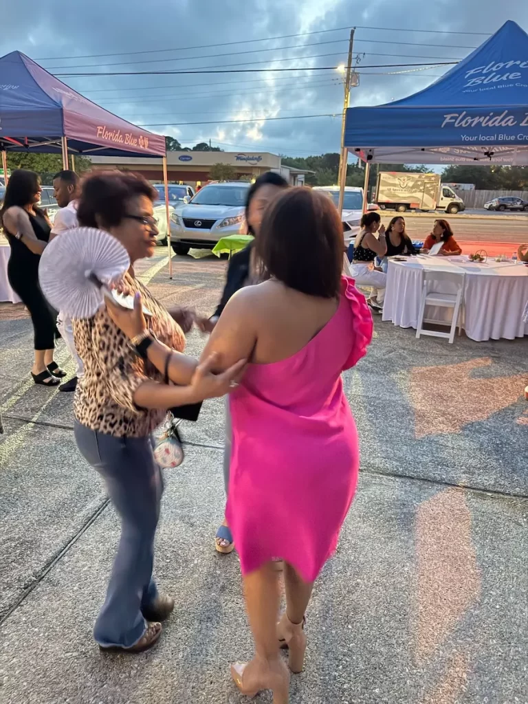 Two women are dancing together outdoors near tables and tents. One wears a pink dress and heels, while the other wears blue pants and a leopard print top, holding a fan. Other people are seated at tables under blue tents in the background.