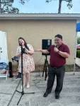 Una mujer con un vestido floral y un hombre con una camisa granate están afuera, junto a unos micrófonos. El hombre habla y gesticula, mientras la mujer sonríe. Están frente a un edificio color canela con altavoces y equipos cerca.