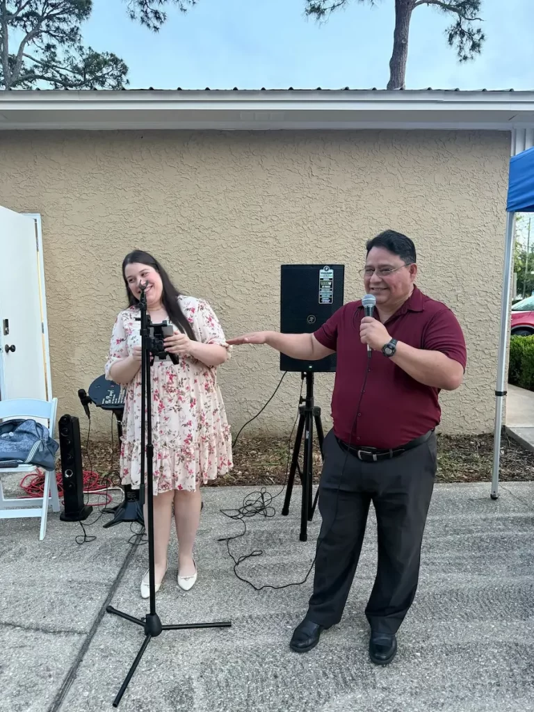 A woman in a floral dress and a man in a maroon shirt are smiling and speaking into microphones outdoors, standing next to each other under a blue canopy by a beige building.