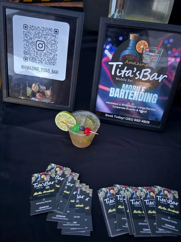 A mobile bartending display with a QR code, promotional signs for "Amazing Tita’s Bar," a cocktail with lime, and business cards on a black tablecloth.
