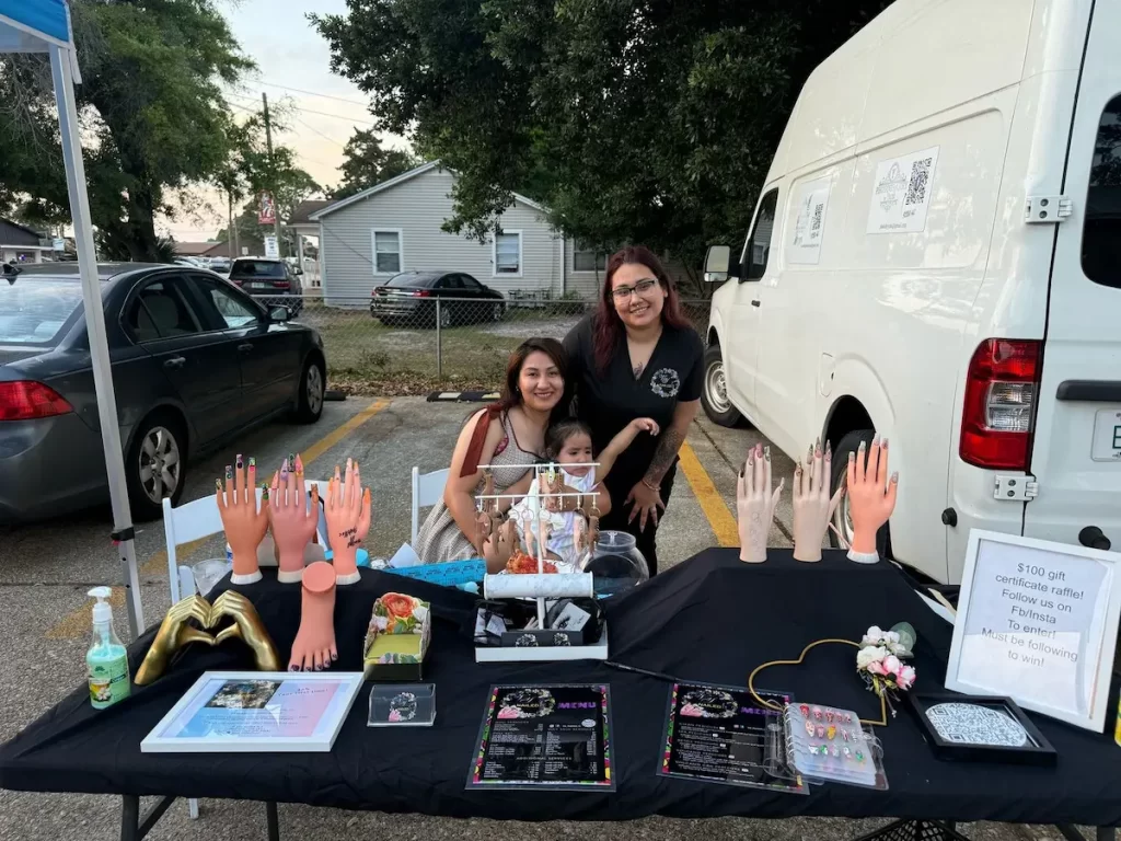 Three women, one holding a small child, stand behind an outdoor table displaying nail art samples, jewelry, and manicure tools. A white van is parked behind them, and a house and trees are visible in the background.