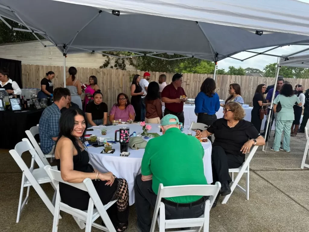 A group of people gathers under canopy tents at an outdoor event. Some are sitting at round tables with white chairs, eating and talking, while others stand and socialize nearby. Food and drinks are visible on the tables.