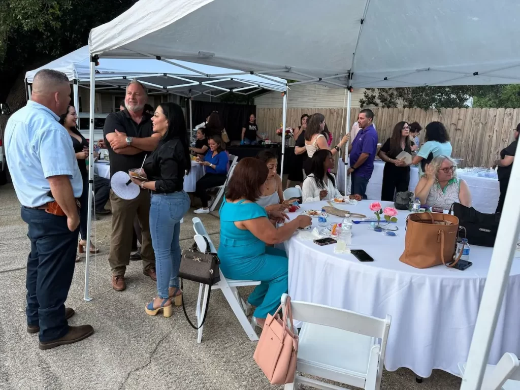 People gather under white canopy tents at an outdoor event, some standing and talking while others sit at round tables eating and socializing. The atmosphere appears relaxed and festive.