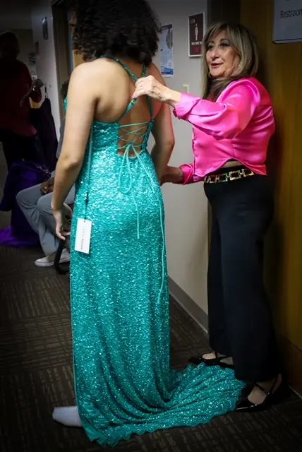 A woman in a sparkly teal dress, part of the 4EVR DEZ team, is being helped with the back laces by another woman in a bright pink blouse and black pants, standing in a hallway with a restroom sign on the door.