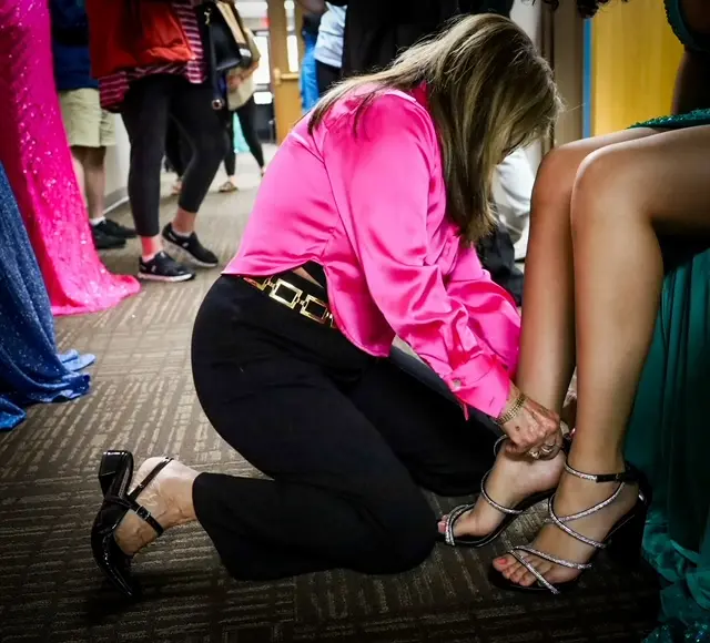 A woman in a bright pink blouse kneels on the floor, helping fasten the silver high-heeled sandal of another woman in a green dress—a quiet moment of compassionate outreach amid people in colorful attire in the background.