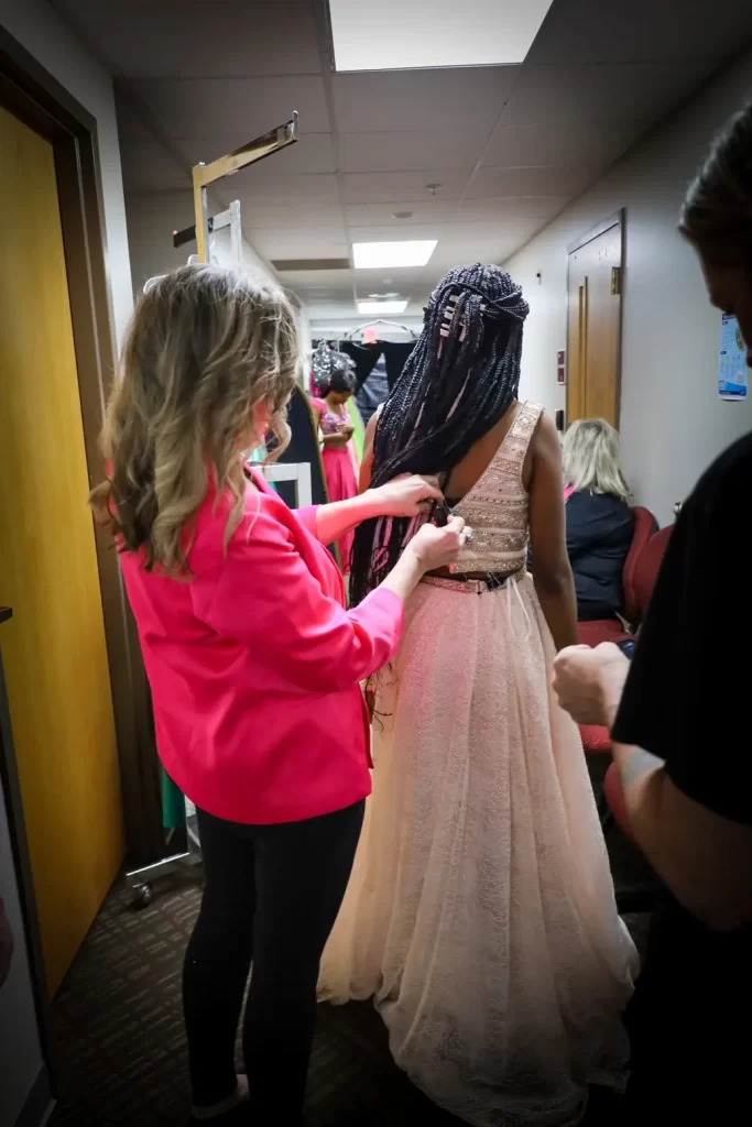 A woman in a pink jacket helps adjust the back of a young Northwest Florida student's elegant, sleeveless, cream-colored gown in a hallway, as other people sit and stand nearby.