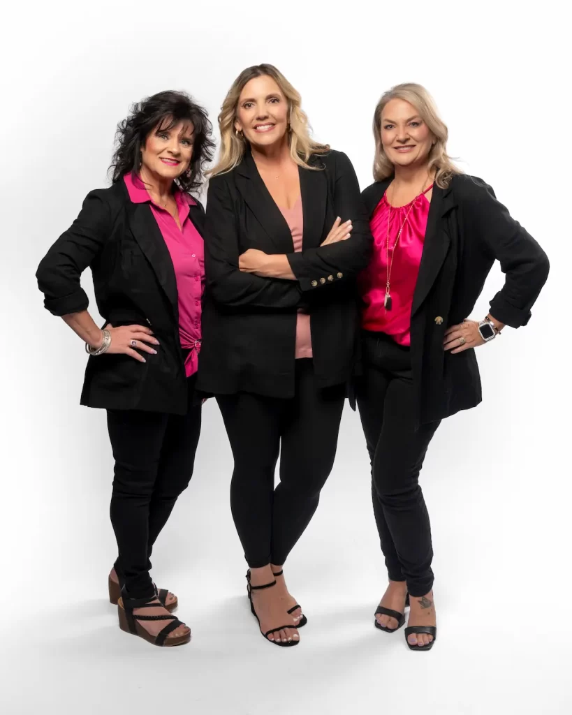 Three women stand together smiling, wearing black blazers and pants with bright pink tops and heeled sandals, against a white background. They pose confidently, embodying 4EVR DEZ's compassionate outreach to Northwest Florida students.
