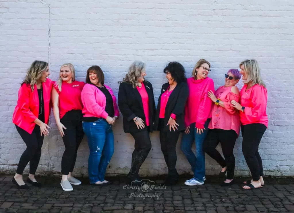 Eight women stand in a row by a white brick wall, laughing and smiling. Most wear bright pink tops with 4EVR DEZ logos, while two wear black jackets. They appear cheerful and engaged in compassionate outreach with each other.