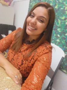 A woman with straight, light brown hair smiles at the camera. She is wearing an orange lace dress, sitting at a table with a gold tablecloth, and there is a leafy green backdrop behind her.