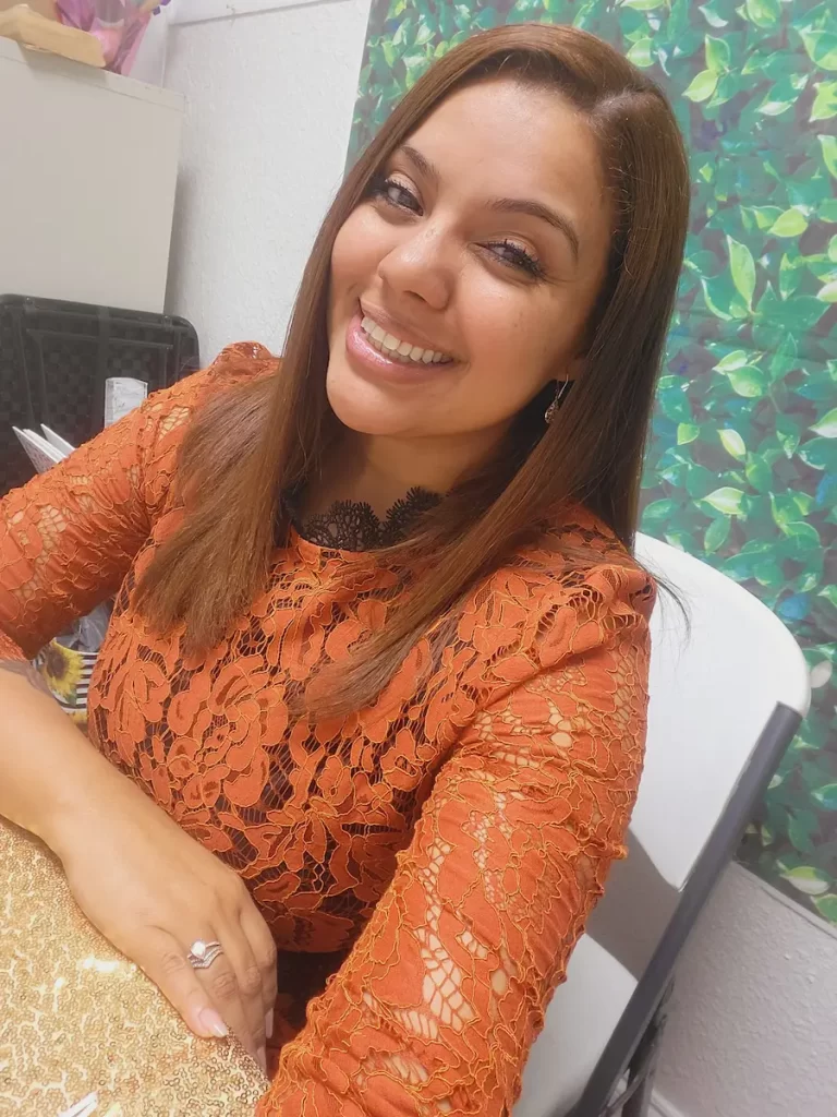 A woman with straight, light brown hair smiles at the camera. She is wearing an orange lace dress, sitting at a table with a gold tablecloth, and there is a leafy green backdrop behind her.