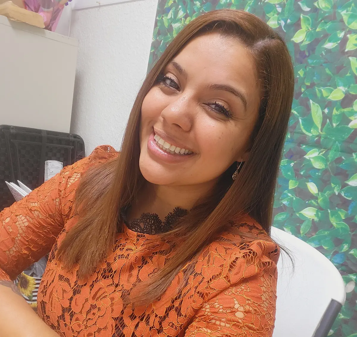 Una mujer de cabello castaño y liso, con un top de encaje naranja, está sentada en un escritorio y sonríe cálidamente a la cámara. Detrás de ella se ve un fondo verde frondoso.