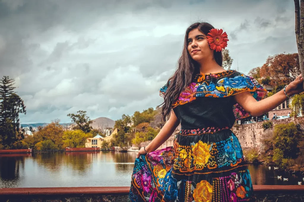 A woman in a colorful traditional dress with floral patterns and a red flower in her hair stands by a lake, celebrating Women’s Health Month. Trees, water, and a cloudy sky form the scenic background.