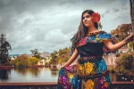 Una mujer con un colorido vestido tradicional con estampados florales y una flor roja en el pelo está de pie junto a un lago, sosteniendo su falda. Al fondo se ven árboles, agua y un cielo nublado.