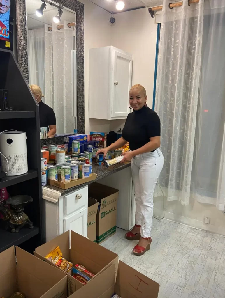 A woman smiles while standing at a kitchen counter organizing canned goods and food for the ONE ANGEL FOUNDATION. Boxes filled with groceries are on the floor nearby. The space is bright with white curtains and cabinets.