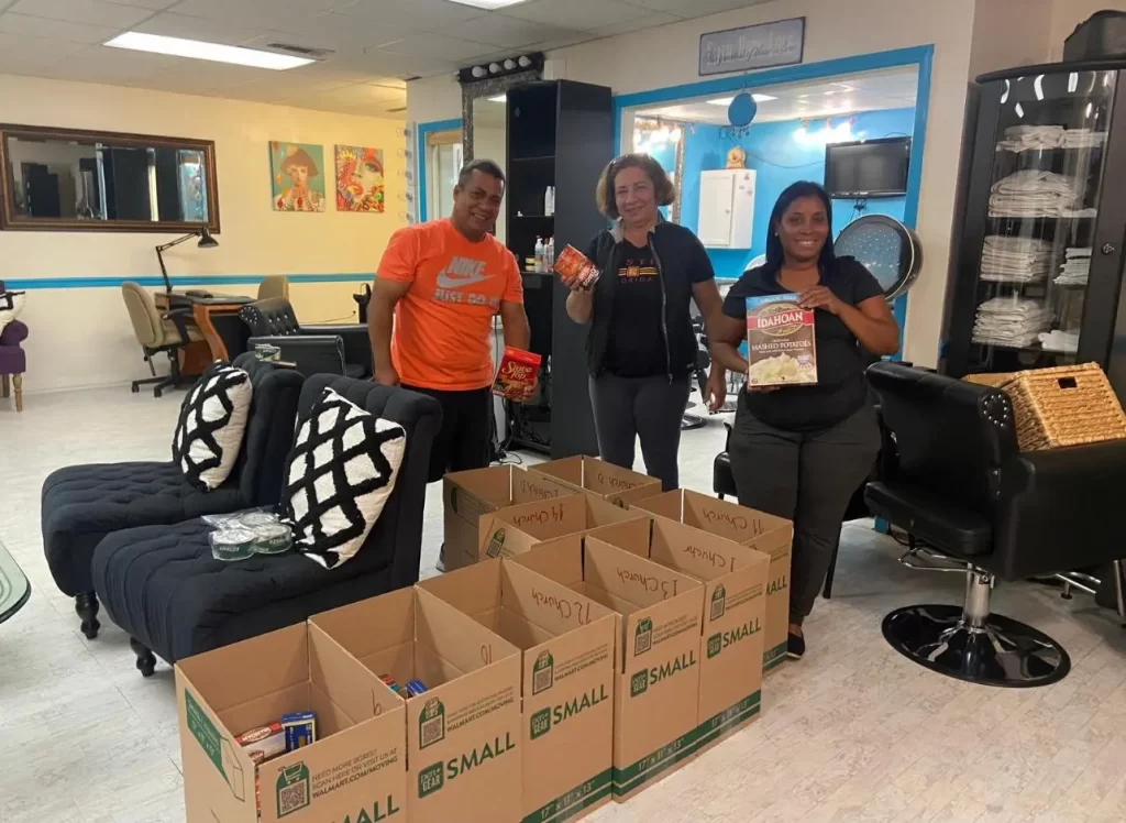 Three people stand in a salon, smiling and holding food items. Several boxes labeled "food" from ONE ANGEL FOUNDATION are filled with groceries on the floor. Salon chairs and equipment are visible in the background.