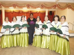 Ocho mujeres con blusas blancas tradicionales a juego y faldas verdes largas con abanicos verdes se encuentran en fila, con una mujer sonriente vestida de negro en el centro, posando en el interior frente a un escenario con cortinas rojas y doradas.