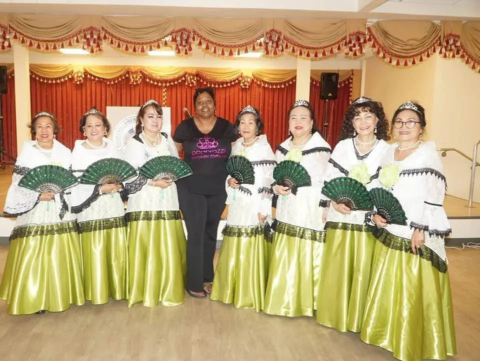 Ocho mujeres con blusas blancas tradicionales a juego y faldas verdes largas con abanicos verdes se encuentran en fila, con una mujer sonriente vestida de negro en el centro, posando en el interior frente a un escenario con cortinas rojas y doradas.