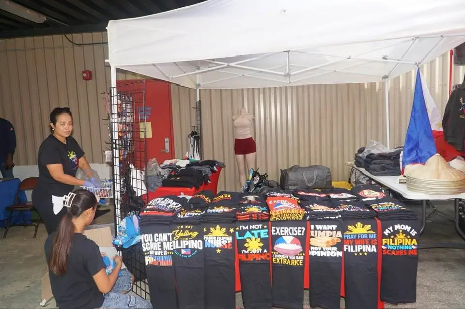 At the Pensacola Asian Pacific American Festival, two women set up a vendor stall with colorful T-shirts featuring Filipino pride slogans and graphics. The shirts line a table under a white canopy, with a Philippine flag visible on the right.