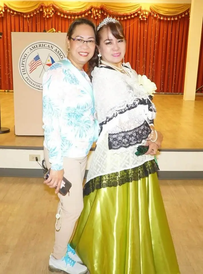 Two women pose and smile indoors at the Asian Pacific American Festival in Pensacola; one wears casual clothes and sneakers, while the other is in a white shawl, tiara, and a shiny green skirt, standing before a stage with a red curtain and podium.