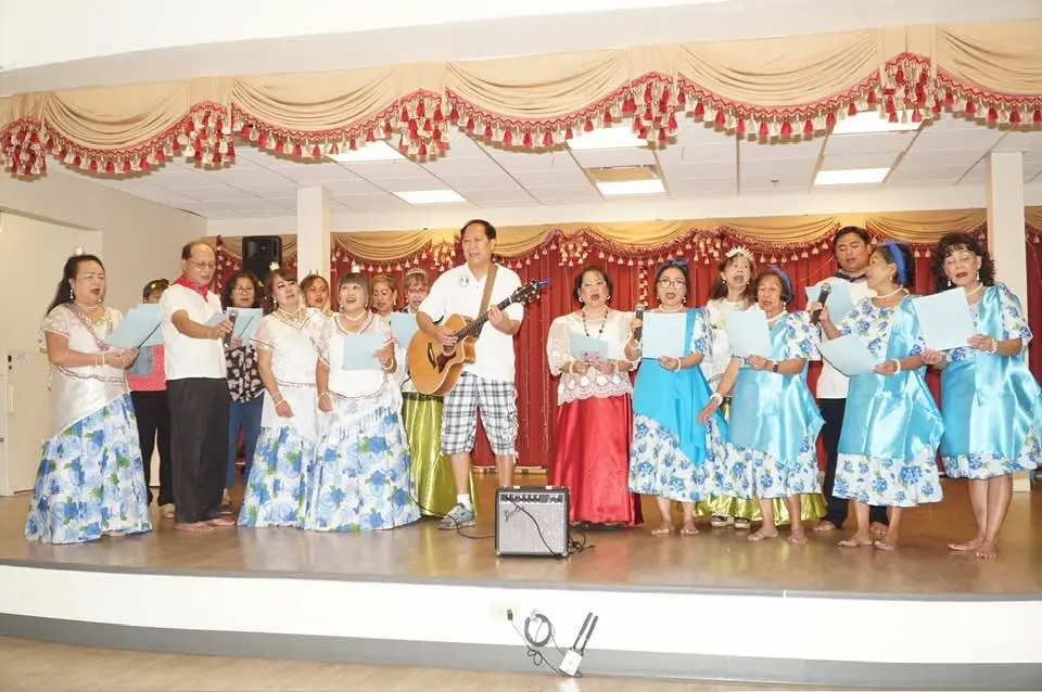 A group of people in colorful traditional outfits stand on a stage at the Asian Pacific American Festival in Pensacola; one man plays guitar while others sing and hold papers. The stage is decorated with red and gold drapes.