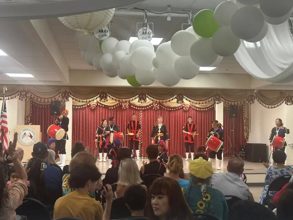At the Asian Pacific American Festival in Pensacola, a group of performers in traditional attire plays drums on a decorated indoor stage. Balloons hang from the ceiling as the audience watches, with curtains and an American flag in the background.