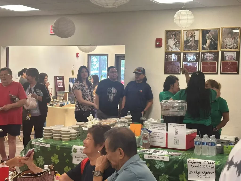 People gather at a community event in a bright room during the Asian Pacific American Festival, with tables displaying food, drinks, and raffle baskets. Some attendees stand and talk, while others sit at a table in the foreground. Decorations and plaques are on the walls.