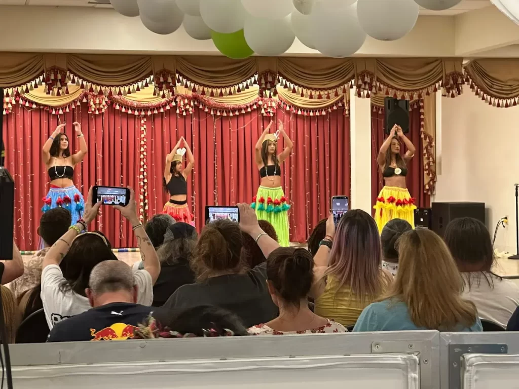 Four women in colorful hula skirts and black tops perform a dance on stage at the Asian Pacific American Festival in Pensacola. Audience members record the lively IL-AM performance, while decorative garlands and white balloons hang above.