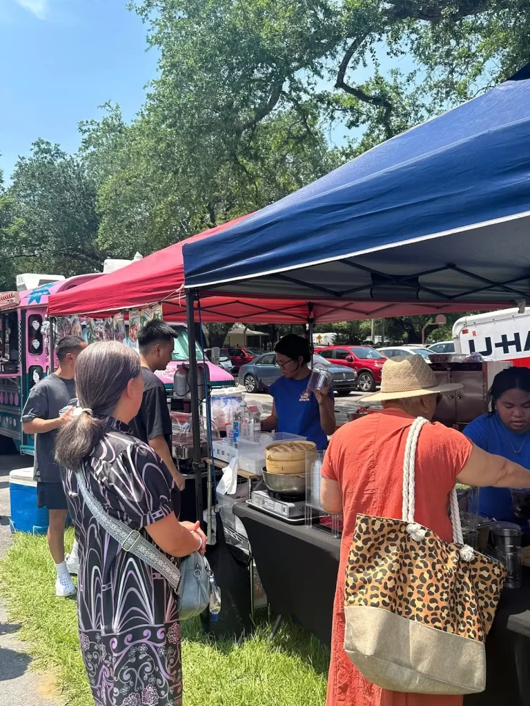 People wait in line under colorful tents at an outdoor food stand during the Asian Pacific American Festival in Pensacola. Drinks, ingredients, and equipment fill the tables, while trees, cars, and other food trucks dot the sunny IL-AM background.