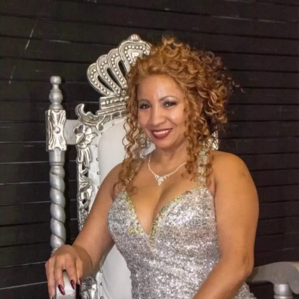 A woman with curly, light brown hair and a sparkly silver dress sits on an ornate silver chair with a crown-shaped back, smiling for the camera against a dark background, proudly representing the Class of 2025.