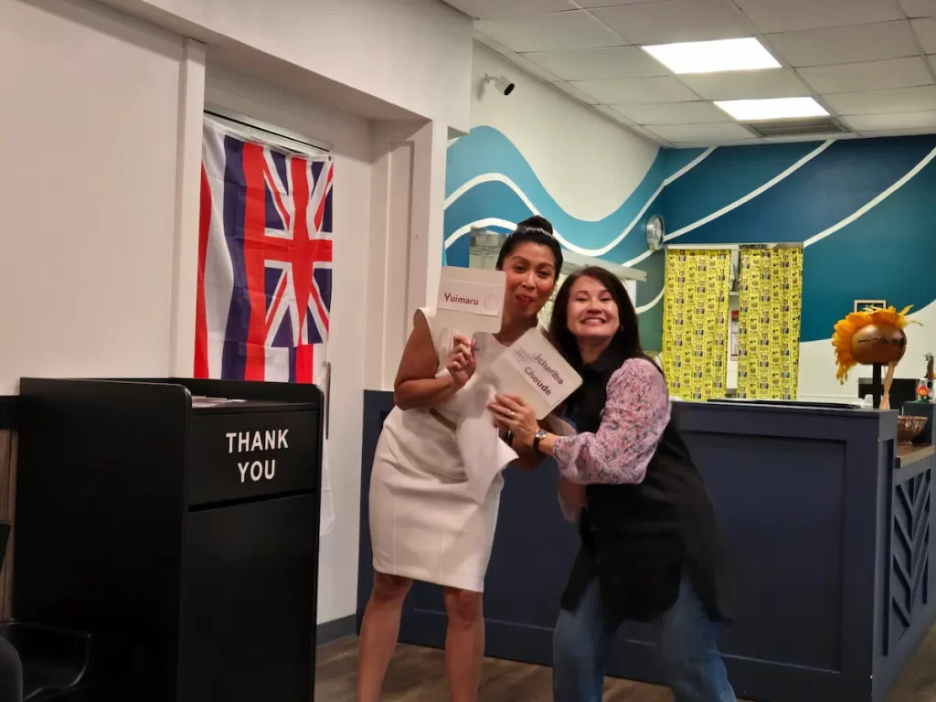 Two women smile and pose indoors, each holding a certificate. A British flag hangs on the wall behind them, and a "THANK YOU" box is visible in the foreground. The setting appears to be an office or reception area.