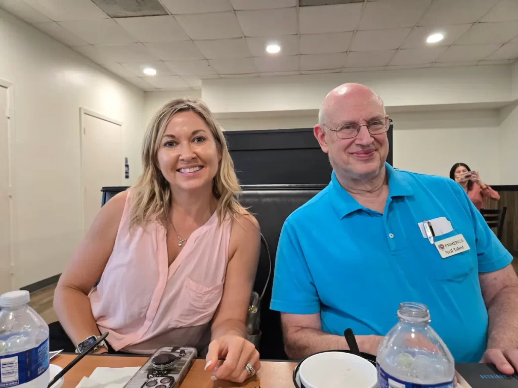 A woman in a pink sleeveless top and a man in a blue polo shirt with a name tag sit side by side at a table, smiling at the camera. Water bottles and other items are on the table in front of them.