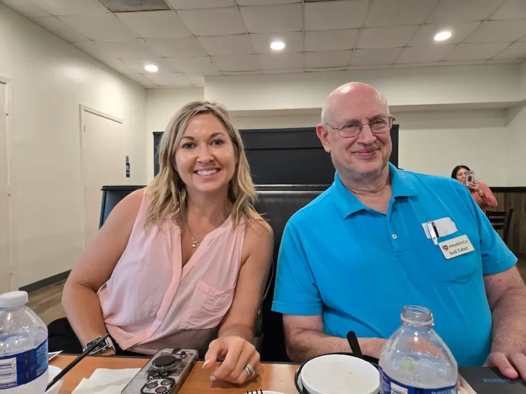 A woman in a pink sleeveless top and a man in a blue polo shirt with a name tag sit smiling at a restaurant table with food, drinks, and utensils. Another person is visible in the background.
