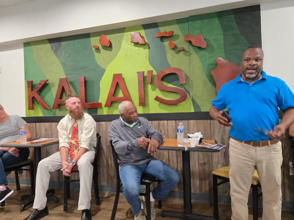 Four men sit and stand in front of a green and red wall sign that reads "KALAI'S." One man in a blue shirt is speaking to the group, while the others listen attentively. They are indoors at a casual gathering.