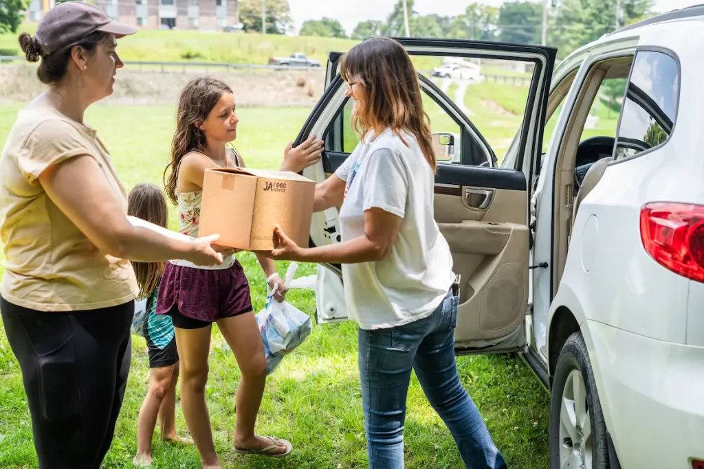 Two women, part of Latino families, stand by an open car door on grass, handing a cardboard box to each other as two young girls wait nearby. A building and trees are visible in the background.