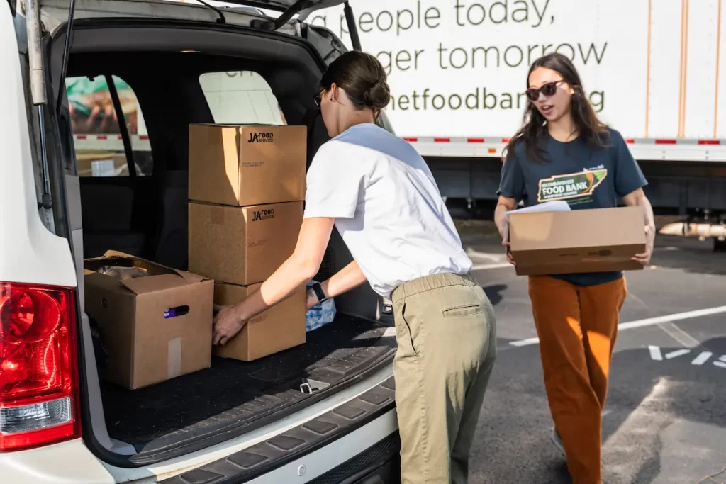 Two women load cardboard boxes into the back of a white vehicle in a parking lot. A truck with No Kid Hungry branding is visible in the background, suggesting food distribution or donation activity.