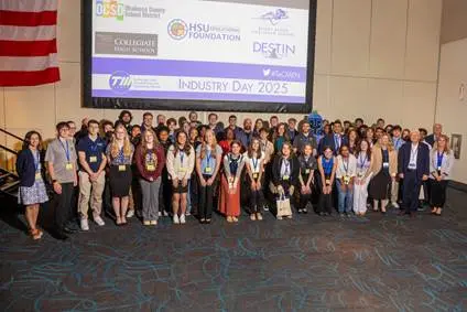 A large group of people pose for a photo in front of a projection screen displaying "TeCMEN Industry Day 2025" and sponsor logos at an indoor event. Okaloosa Students stand among attendees, highlighting local career pathways. An American flag hangs on the left wall.