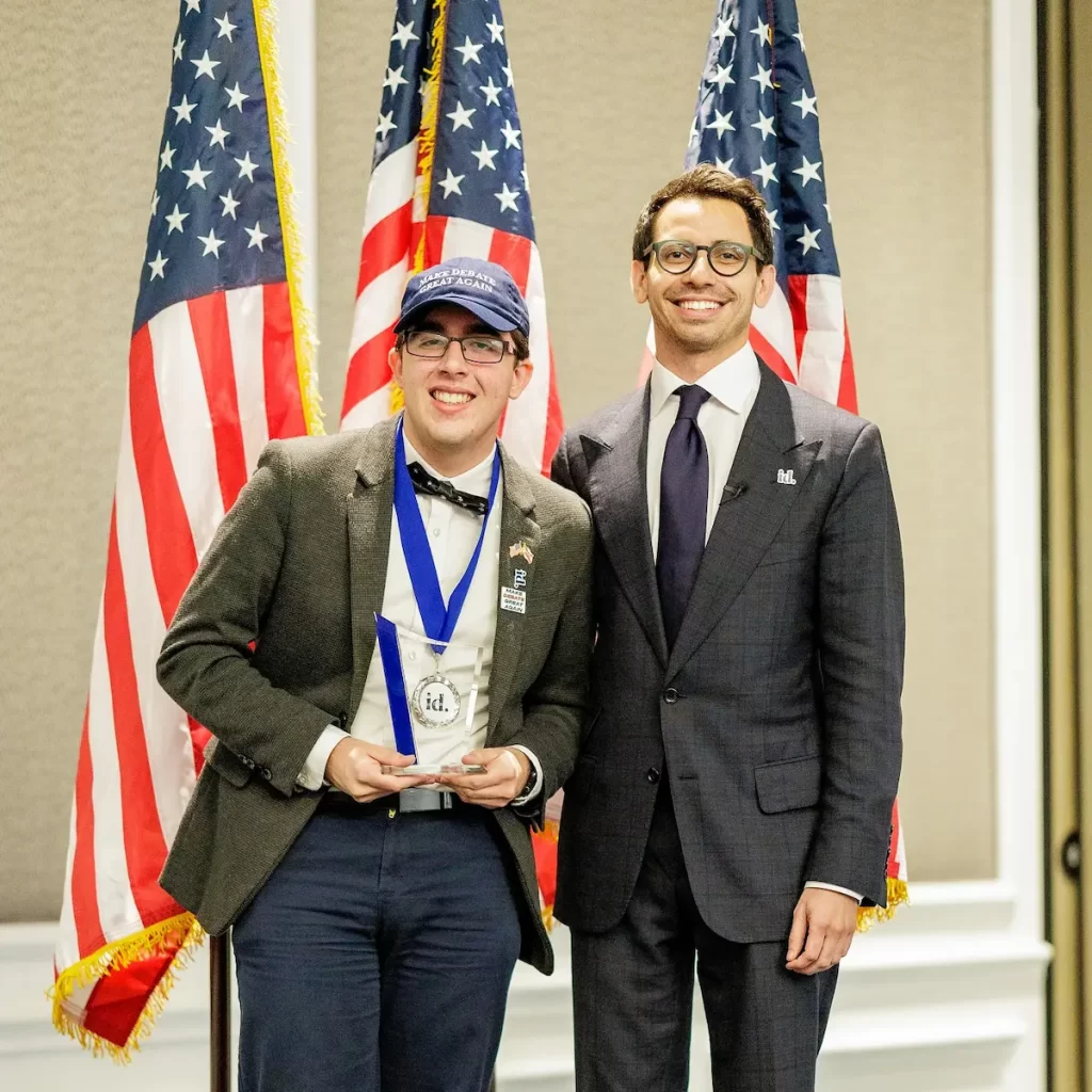 Two men in suits stand smiling before two American flags. William Kiker holds an award and wears a blue medal and hat; the other, in glasses and tie, joins him at what appears to be a National Debate Championship ceremony.