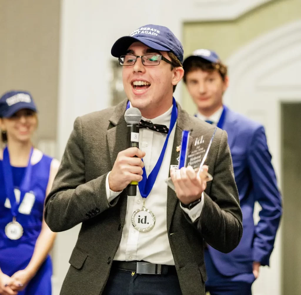 William Kiker, dressed in a suit, bow tie, and cap, smiles while holding a microphone and clear award at the National Debate Championship. Two other people wearing medals and blue attire stand blurred in the background.