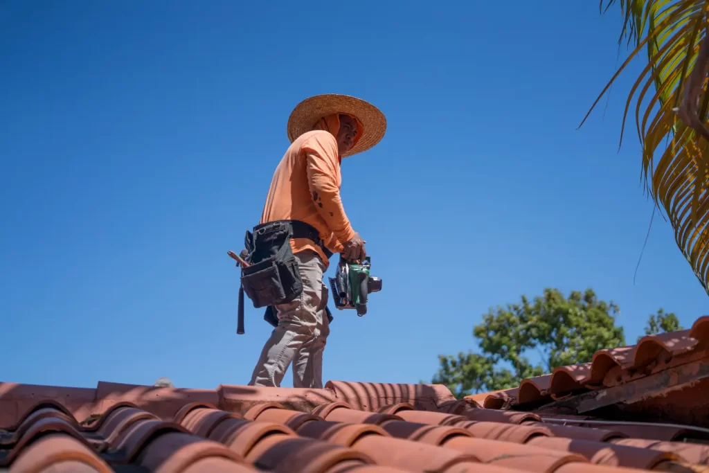 Una persona que viste un sombrero de ala ancha, mangas largas y un cinturón de herramientas se encuentra sobre un techo de tejas en La Pequeña Habana, sosteniendo una herramienta eléctrica bajo un cielo azul claro con hojas de palmeras y árboles visibles cerca.