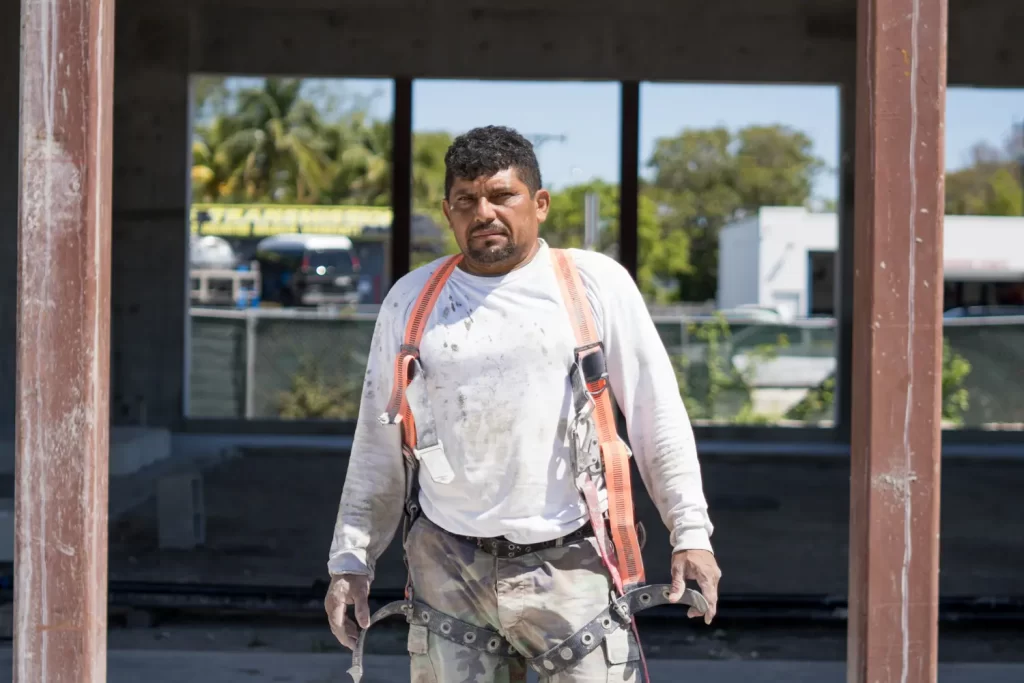 Un trabajador de la construcción que viste una camisa blanca de manga larga, arnés y guantes se encuentra frente a un edificio en construcción en La Pequeña Habana, mirando hacia adelante con una expresión seria.