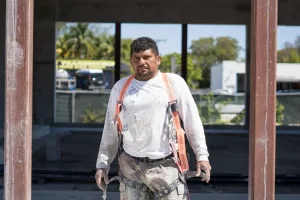 A construction worker wearing a white long-sleeve shirt, safety harness, and gloves stands outside a building under construction in Little Havana, looking forward with a serious expression.