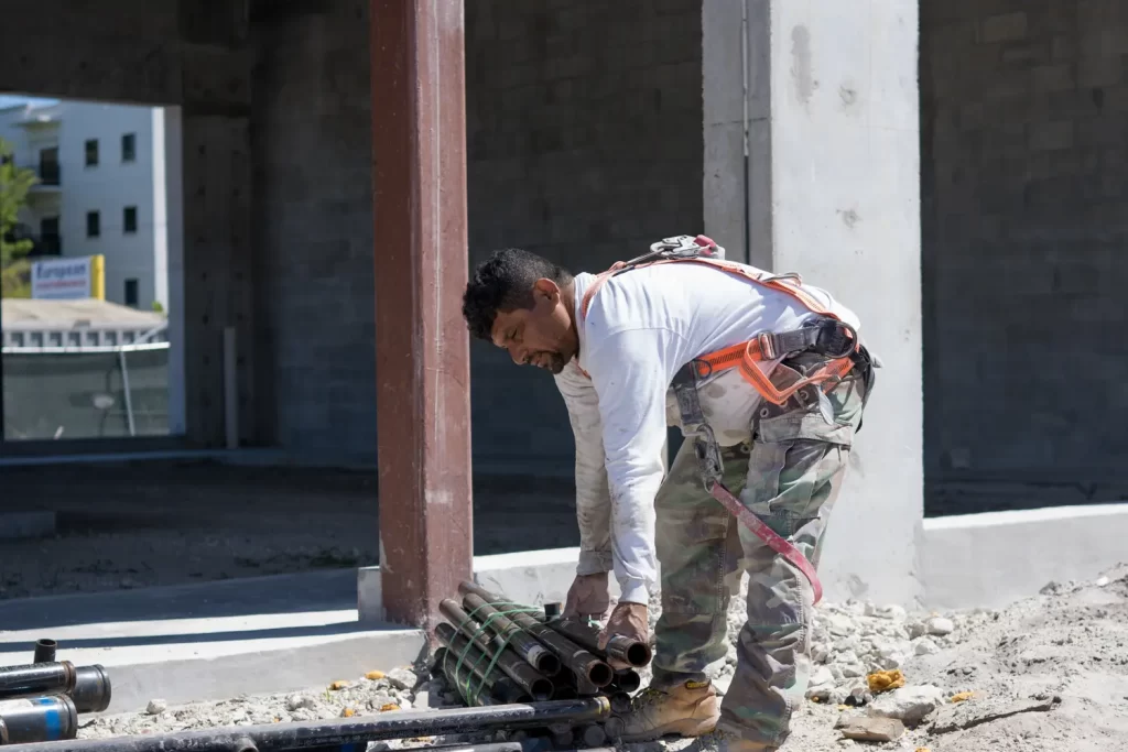 Un trabajador de la construcción que usa un arnés de seguridad y pantalones de camuflaje se inclina para recoger tubos de metal en un sitio de construcción con paredes de concreto sin terminar en La Pequeña Habana.