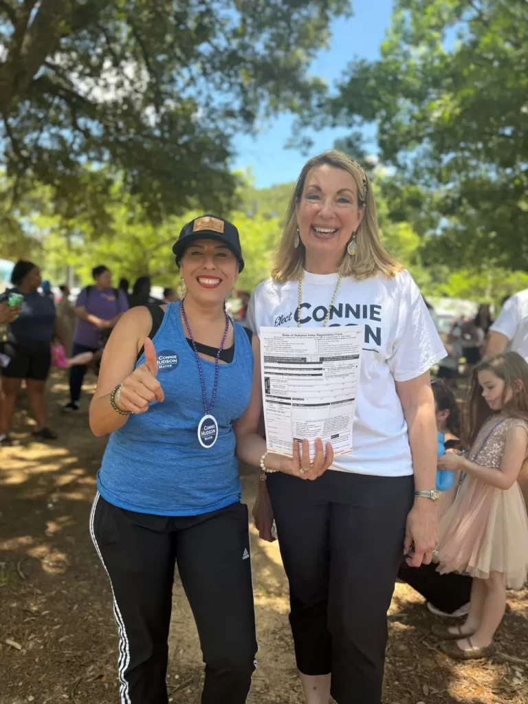 Two women stand smiling outdoors; one gives a thumbs up and the other holds a petition form. People and children are gathered in the background under leafy trees on a sunny day.