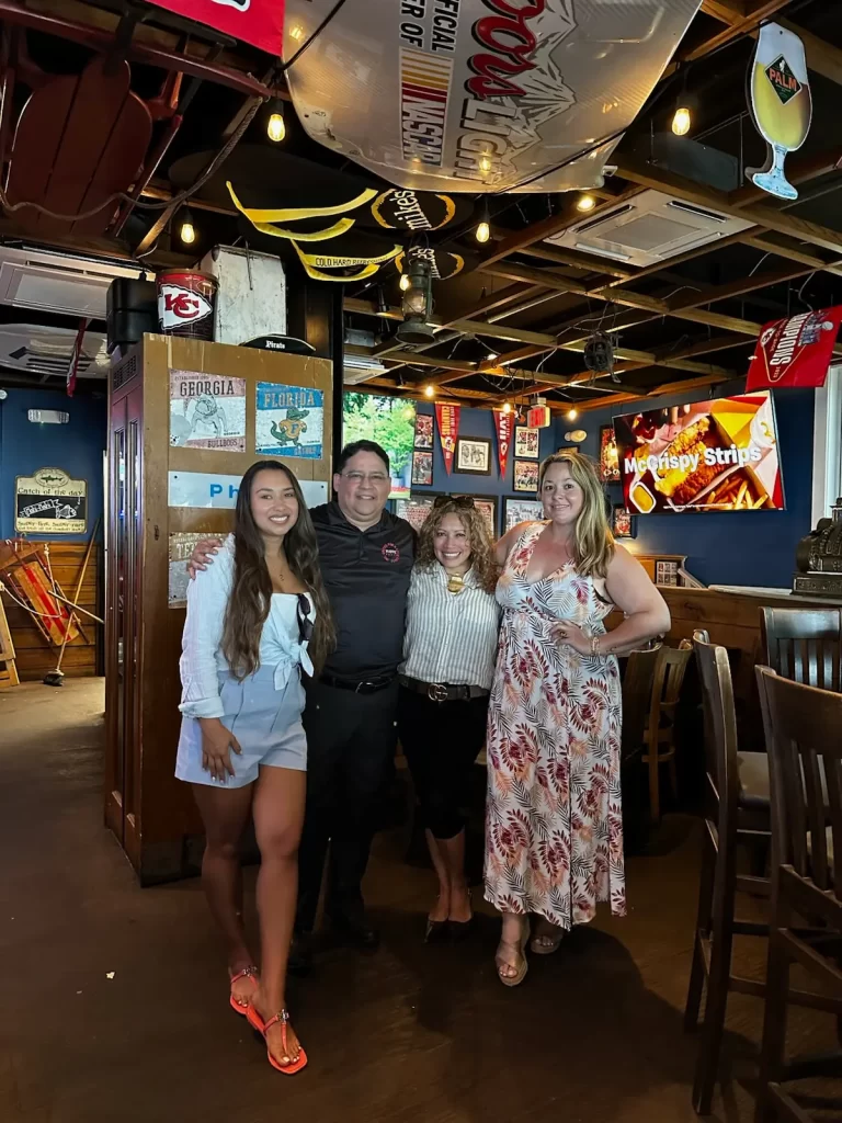 Four people pose and smile inside a sports bar decorated with sports memorabilia, flags, and TVs. Three women are dressed in summer outfits, and one man wears a black uniform. The atmosphere is lively and casual.